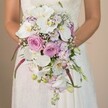 A detailed close-up in Canonbury shows a bride holding a lavish cascading bouquet, its elegant teardrop form brimming with pristine white orchids, blush pink and lavender roses, and ethereal clusters of baby's breath. The arrangement is alive with volume and depth: soft purple stock and sweet pea nestle among the blooms, complemented by dramatic trailing Amaranthus in vivid magenta, whose textured spikes add unique flair. Glossy green foliage and tender buds peek through, balancing classic romance with modern energy. The bride, visible from mid-torso, wears a delicate lace gown, her hands gently cradling the bouquet against the subtle glow of a local interior wall-its neutral tones letting the floral colours sing. The composition feels both timeless and fresh, perfect for a spring or summer wedding near Canonbury's historic gardens. Each element has been thoughtfully arranged, a testament to the skilled floristry found in the neighbourhood and an exquisite inspiration for wedding florals that reflect love stories rooted in the community's charm.