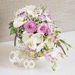 A refined floral composition is shown in a bright, close-up view, perfect for imagining on a wedding table in Canonbury or a romantic celebration near Highbury Fields. The centerpiece is arranged in a round white wicker basket, whose tidy, interlocking weave provides a rustic, handcrafted base for the luxurious flowers above. At the core of the design, full pink roses bloom in generous spirals, each petal curling outward in soft layers that evoke affection and timeless romance. Clustered among the roses are pristine white Phalaenopsis orchids, their broad, graceful petals framing warm yellow and soft amber throats that add a subtle, sophisticated contrast. Ruffled white lisianthus blossoms, some traced with a delicate whisper of lavender along the edges, weave between the larger flowers, creating pockets of airy lightness and visual depth. Slender spires of pale pink stock extend beyond the rim of the basket, gently tumbling outward and bringing a sense of movement, as though the bouquet is spilling across a beautifully laid table. Delicate sprays of white baby's breath float among the main blooms, giving the arrangement a misty, romantic halo that recalls bridal veils and soft evening light. Fresh green foliage is scattered throughout, with slim leaves and emerging buds breaking up the pinks and whites and helping the design feel freshly gathered and alive. In front of the basket, two decorative spheres made from pale wicker or rattan rest on a clean white surface, echoing the basket's texture and enhancing the natural, organic feel. A neutral, softly textured beige backdrop frames the entire scene, allowing the flowers' colours and forms to stand out clearly, ideal for illustrating Canonbury wedding table arrangements, anniversary dinners, or elegant engagement celebrations.
