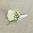 A single white orchid boutonnière is displayed on a pale, sand-coloured fabric with a fine, woven texture, the kind of understated backdrop that allows every floral detail to come forward. The orchid is the star: its large, velvety petals stretch outward in perfect symmetry, pure white at the edges and gently warming into a soft cream near the centre. Within the throat of the bloom, a subtle hint of buttery yellow glows like a captured beam of light, giving the flower a sense of depth and quiet vitality. Surrounding this central orchid, dainty stems of white baby's breath form wisps and clusters, each tiny bloom creating a mist-like frame that adds volume without heaviness, rather like the drifting blossom clouds you might see along New River Walk in spring. Slender strands of fresh green foliage peek out from beneath, some as narrow as blades of grass, others with a slightly more feathery line, adding movement and an echo of garden greenery to the compact design. The stems of the orchid and filler flowers are tightly bound, then wrapped in smooth, luminous white satin ribbon, which forms a clean, cylindrical handle that sits comfortably in the hand before being pinned to a jacket. Just below the orchid's base, a small, neatly tied satin bow punctuates the design, finishing it with a traditional wedding-day flourish. The soft, even lighting accentuates the contrast between the creamy petals, the crisp white baby's breath, and the fresh green foliage, creating a sense of serene anticipation. It feels like a groom's boutonnière prepared by a Canonbury florist for a chic, local celebration, ready to be worn with pride along the leafy streets leading toward Islington's historic squares.