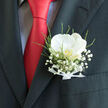 This image shows a classic wedding boutonnière, carefully slipped into the pocket of a charcoal suit jacket, framed by the strong diagonal of a bright red tie. The focal flower is a single white orchid, opening with serene poise, its petals broad, velvety, and gently cupped as if cradling a secret. At its centre, the orchid holds a subtle golden hue, a small sunlit glow that hints at tenderness and joy. Encircling the orchid, clouds of baby's breath form a delicate mist of white, each tiny bloom contributing a sense of softness and airy romance. Fine stems of green foliage and feathery fern provide contrast and texture, mimicking the lush greenery of nearby Canonbury Square gardens, and giving the arrangement a sense of fresh movement, as though a soft breeze had just brushed through. A slim white ribbon at the base, neatly tied and perfectly aligned, secures the stems with an artisan's care, its satiny surface catching the light in small, refined highlights. The overall palette stays crisp and elegant - white, green, charcoal, and a bold ribbon of red from the tie - creating a striking visual harmony. This boutonnière feels ideally suited to a groom or groomsman stepping out for a ceremony in Canonbury or along the quiet streets leading to Islington Town Hall, embodying modern style grounded in timeless floral tradition. The piece suggests the gentle fragrance of fresh blooms, the rustle of a well-fitted suit, and the steady heartbeat of someone about to say I do.
