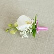 A delicate orchid boutonniere lies horizontally on a softly textured, oat-coloured linen backdrop, the neutral fabric letting every floral detail come forward as if in quiet conversation. At the centre, a single white Phalaenopsis orchid opens in full, its broad petals smooth and velvety, curving gently outward like small porcelain wings. In the heart of the bloom, a touch of soft yellow and the faintest blush of pale pink draw the eye inward, adding warmth and depth without disturbing the overall purity of the flower. Around the base of the orchid, tiny clusters of white baby's breath form a light, cloud-like collar, each minuscule bloom creating a misty, romantic halo that softens the silhouette. Slender green leaves slip out from behind the flowers, their fresh, vibrant tone a natural contrast to the crisp white petals. To one side, two lime-green orchid buds rest on a slim stem, still tightly closed, suggesting new beginnings and blooms yet to come - a fitting symbol for a wedding day or prom evening in Canonbury, whether near the greenery of New River Walk or a townhouse celebration off Essex Road. The stems of the arrangement are gathered neatly and wrapped in a bright fuchsia pink satin ribbon, the bow tied with care so its small loops and tails gleam softly against the linen. The satin's glossy sheen adds a joyful, celebratory note, hinting at laughter, photographs, and the gentle rustle of formal attire. Soft, even lighting glides over the scene, highlighting the subtle textures of petal, ribbon, and fabric, and lending the whole boutonniere an air of calm refinement. This is a quietly elegant floral accessory, the kind a local Canonbury florist might pin to a lapel just before the ceremony begins, capturing a moment of poised anticipation.