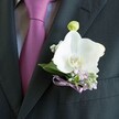 A close-up photograph shows a perfectly tailored charcoal or deep black suit jacket, the fabric smooth and finely woven, with the left lapel brought crisply into focus. Against this dark canvas rests a single white Phalaenopsis orchid boutonniere, its broad, satiny petals opening like wings and catching the soft ambient light, so they almost glow. At the flower's heart, a hint of buttery yellow and warm orange in the stamen adds a gentle touch of colour, echoed by nearby clusters of tiny lavender and pale lilac blossoms that may be heather or another delicate filler. Sprigs of white gypsophila, or baby's breath, thread between the lilac tones, creating a misty halo of texture, while polished deep green leaves frame the orchid and keep the composition balanced. Just above the main bloom, a tightly closed pale green orchid bud points upward, suggesting new beginnings and the quiet anticipation of a wedding morning somewhere in Canonbury, perhaps near the leafy calm of Highbury Fields. A slim satin ribbon in lavender or mauve is tied into a neat bow at the base, securing the stems just above the suit's breast pocket and echoing the nearby tie in rich plum or magenta, which peeks out from a light grey, subtly patterned shirt. The overall mood is refined, romantic, and carefully composed, the sort of boutonniere a local Canonbury groom might wear for a ceremony in an elegant townhouse or a small Islington venue-understated but full of meaning, with every petal chosen to convey poise, affection, and a sense of thoughtful celebration.