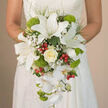 The image captures a bride from the waist down, her hands gently wrapped around a luxurious trailing bouquet that rests against the soft, off-white lace of her wedding dress. The arrangement is composed in a refined teardrop shape, beginning with several stately white oriental lilies whose wide, velvety petals are marked with delicate reddish-brown speckles and long, graceful stamens. Just beneath her fingers, a perfectly formed ivory rose sits as the romantic focal point, its creamy swirls of petals framed by tiny, cloudlike tufts of white baby's breath. From this central cluster, a waterfall of white Phalaenopsis orchids cascades elegantly, each bloom arching downward to create a sense of motion, like petals drifting along the New River Walk in Canonbury on a still morning. Punctuating the white are bright lime-green, mossy-looking balls of Green Trick dianthus or pompon chrysanthemums, their fuzzy texture and vivid colour adding a modern, playful edge. Bright red hypericum berries are threaded throughout the bouquet, small but striking accents that lend depth and a gentle festive note. Fine sprigs of feathery greenery and darker, glossy leaves peek out at the edges, framing the composition and enhancing its layered, sculpted appearance. The lighting feels soft and natural, highlighting every curve of petal and vein of foliage, as though the bouquet has just been delivered fresh from a local Canonbury florist for a ceremony in a nearby townhouse or church. The overall effect is one of poised elegance, freshness, and heartfelt celebration.