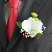 A close-up wedding portrait focuses tightly on a single elegant boutonniere pinned to the lapel of a dark charcoal suit jacket, its fine woven texture softly catching the light. At the centre of the design, a flawless white Phalaenopsis orchid opens like a pair of wings, its smooth petals gently curved and its yellow-and-white lip adding a small, sunlit detail. Tucked around the orchid are sprigs of airy white baby's breath, each tiny bloom floating like mist against the deep fabric, and two bright lime-green chrysanthemum button blooms that bring an unexpectedly playful pop of colour. Glossy green leaves cradle the flowers, framing them in natural freshness. Just beside the boutonniere, a vivid red satin tie lies neatly over a light grey and white small-checked dress shirt, the tie's sheen mirroring the slim, deep red ribbon that wraps and secures the flower stems at the base. The composition feels intimate and thoughtful, as though captured moments before a Canonbury wedding ceremony near the quiet streets off Essex Road, with the groom taking a final breath before stepping out. The contrast between the crisp tailoring, the sculptural orchid, and the delicate cloud of baby's breath speaks of careful craftsmanship and calm confidence. You can almost imagine this fresh, cool corsage being pinned on in a townhouse just off Canonbury Square, a small but meaningful flourish prepared by a local florist who understands how a single flower detail can complete a wedding look and carry the emotion of the day.