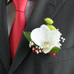 A finely tailored dark suit lapel, likely chosen for a Canonbury wedding celebration, is adorned with a thoughtfully crafted boutonnière that blends classic style with contemporary finesse. At its heart, a single orchid in pristine white unfurls gently curved petals, their surface silky and luminous, with a soft cream-yellow flush at the centre that draws the eye in. Behind this focal bloom, two lime-green button chrysanthemums nestle close, their spherical shapes and tightly set petals adding texture and a lively pop of colour, like fresh shoots emerging in nearby New River Walk gardens. Around them, small sprays of baby's breath create a delicate halo, each tiny white flower floating on fine stems that soften the edges of the design. Slender leaves in a deep, glossy green weave discreetly through the composition, adding structure and a subtle sense of movement, as if the flowers have just been caught in a gentle breeze drifting across Canonbury Place. The base of the stems is neatly bound with a narrow red ribbon, rich and velvety in tone, which mirrors the sheen of the groom's deep red tie, tying the look together with a romantic, almost vintage charm. The contrast between the crisp white orchid, lively green chrysanthemums, and the dark, matte fabric of the suit creates a harmonious balance-neither too bold nor too faint, but quietly confident. This boutonnière feels especially suited to intimate ceremonies and receptions, a graceful floral accent that accompanies its wearer from formal photographs along Upper Street to late-night toasts, always suggesting care, intention, and a touch of local Canonbury elegance.