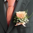 A classic wedding buttonhole rests elegantly on a gentleman's lapel-a quietly stunning arrangement of fresh garden blooms created for the heart of Canonbury. At its center is a single rose in creamy peach blush, its petals opening softly and offering a subtle radiance reminiscent of first light over the Regent's Canal. Encircling the rose, delicate sprays of baby's breath introduce a frothy, cloudlike lightness, while slender arcs of greenery and a gentle dark leaf bring natural freshness and depth. The boutonniere sits against the structured charcoal of a perfectly tailored suit, complemented by a peach satin bow at the base, echoing the lively tone of the neatly tied neckwear. The effect is both polished and tranquil, suitable for a local bridegroom or prom attendee awaiting photographs near Canonbury's iconic pubs and leafy squares. Crafted by local hands, it's more than adornment-it is a subtle promise, a fragrant whisper of garden mornings, unity, and celebration, ready to accompany cherished moments along the tree-lined streets of N1.