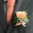 Pinned low on a charcoal suit lapel, a single peachy-pink rose becomes the quiet centre of attention, its petals layered with the kind of softness that suggests it was chosen at the peak of freshness. The blossom's warm blush is set off by light, feathery strands of green foliage and clusters of tiny white baby's breath-gypsophila-that float above the rose as if breathing around it. At the boutonnière's base, a slim satin ribbon in a pale salmon tone is tied with care, mirroring the sheen of a coordinating tie and contrasting gently with the subtle dark grid of a pale shirt beneath. The close-up framing highlights texture: the rose's velvety curls, the airy lace of gypsophila, the fine serration of the greenery, and the smooth matte of the suit fabric. Soft daylight washes the scene, lending a natural warmth to the peach hues and creating a sense of calm anticipation-an accessory made to be worn at a wedding, an anniversary, or a formal celebration. The arrangement's asymmetry and hand-tied finish speak to artisanal craft, where each tiny filler stem is placed to enhance the bloom rather than overwhelm it. There is, too, a local intimacy to the image: one can picture this boutonnière arriving from a nearby florist, prepared for an aisle walk past Canonbury Square or a reception on a quiet road off Essex Road, bringing a whisper of garden-sweet fragrance to the day's gentle rituals.