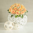 A richly textured study in soft apricot and cream, this arrangement gathers peach roses into a generous, domed cluster that reads as both plush and restrained. Each rose opens with a gentle gradient-from a pale, creamy centre to a warm, honeyed apricot edge-inviting a fingertip to follow the curl of each petal. Around and between those roses, clouds of white gypsophila add an ethereal veil, the tiny star-like flowers catching light like scattered pearls and lending an airy lift that prevents the mass of blooms from feeling heavy. The whole sits within a sculptural white woven orb, its organic rattan structure visible beneath and around the flowers, the weave creating a sense of movement and modern craft. Leaves of bright green peek through, their matte surfaces grounding the composition and adding crisp contrast to the soft blooms. Light in the photograph is soft and directional-morning or late-afternoon sun-that highlights petal texture and creates delicate shadow play across the wicker. The scent is imagined as powdery and sweet, a light reminder of fresh-cut roses and summer hedgerow. This is an arrangement suited to weddings, anniversary gifts, or an elegant housewarming for a Canonbury townhouse near Essex Road or St. Paul's Road; it feels handcrafted by someone who knows local interiors and simple, refined taste. Prepared in a small Canonbury workshop, the piece reflects close attention to balance, rhythm, and the tactile joy of working with fine, seasonal roses and airy gypsophila to produce an object that calms as much as it celebrates.