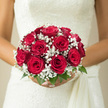 In this image, a bride stands poised in her wedding dress, holding a perfectly rounded bouquet of red roses and white baby's breath that seems to glow with quiet intensity. The roses are a true, romantic red-neither too bright nor too dark-each bloom fully open yet still structured, with overlapping petals that look almost like folded velvet. They are grouped in a tight, circular cluster that fills the hands, creating a sense of fullness and stability, like a small, living jewel box. Threaded between these passionate blooms are airy sprigs of gypsophila, their tiny white blossoms forming a soft, frothy border that lightens the whole design. The baby's breath appears almost weightless, reminiscent of a fine, early-morning mist drifting over the quiet streets near Canonbury Square, lending an ethereal charm that softens the drama of the red. A gentle play of light catches on the rose petals and the minute flowers, bringing out subtle variations in tone and texture, from satin-smooth petals to the crisp, almost papery florets of gypsophila. A slim collar of fresh green leaves anchors the bouquet at the base, giving just a hint of foliage without overwhelming the refined shape. The stems are bound tightly with pale ribbon to match the bridal gown, suggesting readiness for a long day of celebrations, perhaps moving from a Canonbury townhouse ceremony to a reception nearby in Islington. The overall effect is one of romantic tradition-a bouquet that feels intimate, heartfelt, and perfectly suited to being held in photographs and memories long after the last guest has gone home.