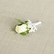 A single white rose boutonniere lies diagonally across a softly textured, light beige fabric that looks like natural linen, its fine weave running in gentle diagonal lines under the flowers. At the heart of the design is a creamy white rosebud, petals still tightly furled yet full and velvety, suggesting a bloom chosen at just the right moment before opening. Tiny hints of green sepals cradle the base of the rose, giving a fresh, newly cut feel. Around the rose, delicate tufts of white baby's breath form airy clouds of miniature blossoms, each floret no bigger than a pinhead, adding a light, romantic halo. Slender, deep green leaves and a few slightly feathery sprigs of foliage frame the flowers, the greens clean and vibrant, offering a cool contrast to the warm cream tones. The stems are gathered neatly and wrapped in smooth, pristine white satin ribbon, finished with a small, carefully tied bow that sits just below the bloom, signalling that the piece is ready to be pinned to a lapel for a wedding or formal evening on the quiet streets of Canonbury. The lighting is soft and even, as though filtered daylight from a nearby townhouse window, gently highlighting the subtle curves of the rose petals, the tiny spheres of baby's breath, and the sheen of the ribbon. The overall mood is calm and elegant, evoking the anticipation of a groom or groomsman preparing for a ceremony in a nearby Canonbury garden or along the tree-lined paths that connect to Highbury Fields, where classic, understated flowers like this rose boutonniere feel perfectly at home.