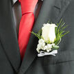 This close-up shows a timeless wedding boutonniere, the kind of thoughtful finishing touch often seen at Canonbury receptions and intimate gatherings along Upper Street. A single white rose sits at the heart of the design, its petals gently unfurling in natural spirals, creamy and soft-edged, with just enough openness to suggest a day in full bloom. The rose rests against a charcoal suit lapel, the contrast between the pale flower and dark cloth creating a clear focal point that draws the eye without overpowering the overall look. Surrounding its base, clusters of snow-white baby's breath form a fine, airy frame, each tiny blossom like a speck of light, adding texture and a sense of delicacy. Slender tendrils of deep green foliage loop and curl around the rose, while feathery ferns extend a little beyond the pocket, evoking the gentle greenery of Canonbury's garden squares on a quiet morning. At the stem, a neatly tied white ribbon bow holds everything in place, the satin looking smooth and cool to the touch, giving a subtle nod to classic menswear styling. The arrangement feels ceremonial and composed, right at home at a wedding, prom, or formal event where understated grace is appreciated. The lighting suggests a natural, soft brightness, like daylight filtering through a townhouse window, catching the rose's velvety texture and the suit's fine weave. The boutonniere sits securely in the breast pocket, poised for photographs, handshakes, and embraces that will be remembered long after the petals have faded.