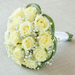 There is a dreamlike calm to this bridal bouquet, composed of roughly fifteen to twenty soft, creamy-yellow roses clustered into a generous, rounded dome. Each rose displays a perfect spiral of petals, velvety to the eye and subtly luminous under soft, diffused light. Scattered between the larger blooms are delicate sprays of white Gypsophila that create an airy, cloud-like halo, lending lightness and innocence to the composition. Around the edges, long, slender arcs of vibrant green leaf - probably lily or bear grass - curve with an elegant, natural motion, framing the roses and suggesting the gentle sway of a summer garden. The stems are neatly exposed at the bottom and wrapped in pristine white satin ribbon, finished with a tight knot that speaks of careful hand-tying; the handle would feel cool and reassuring to hold during a procession down Canonbury Road or across the lawn at Highbury Fields. The bouquet sits on a soft taupe linen cloth that complements the pale hues and lets the subtle vanilla-tinged cream of the roses take centre stage. The overall impression is one of serene celebration: a scent of clean rose tempered by green leaf, a tactile mix of velvety petals and fine baby's breath, and a balanced silhouette that reads as both traditional and quietly modern. It's a composition I would recommend for weddings, vow renewals, or intimate anniversary moments around Islington and the surrounding neighbourhoods.