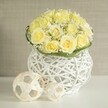 A serene, contemporary floral centerpiece is arranged with the care of an experienced Canonbury wedding florist, blending romance with clean design. Dominating the composition is a lush dome of soft, pale yellow roses, each bloom unfurled to reveal layers of velvety petals in tones of fresh cream and warm butter. They nestle close together so that the overall impression is one of fullness and luxury, without appearing heavy. Tucked among the roses, stems of white baby's breath introduce a fine, frothy texture; their minute blossoms scatter light like tiny stars, creating a gentle contrast that softens the structure. At the base of the bouquet, a neatly wrapped band of green foliage, perhaps woven leaves or slender blades of grass, encircles the stems, adding a crisp, botanical edge that quietly frames the flowers.
Supporting this floral dome is a distinctive white woven vase, sculpted from smooth strands that crisscross and loop in an open, basket-like pattern. The vessel's shape subtly echoes a modern urn, broadening slightly at the top, narrowing at the centre, then widening again towards the base. Its airy lattice allows delicate shadows to play across the bright white tabletop below, reminiscent of dappled light under the trees near Canonbury Gardens. To one side, two decorative spheres crafted from pale, natural-looking strips of wood or rattan continue the theme of open weaving, their different sizes introducing a sense of gentle movement and balance. The larger sphere sits closer to the viewer, with the smaller tucked just behind, like art objects placed carefully for a wedding reception or intimate dinner party. The background is a softly blurred wash of warm beige or light grey, with a subtle texture that never competes with the main display. Overall, the image conveys quiet celebration and timeless elegance, suitable for sophisticated wedding tables, engagement parties, or refined home decor across Canonbury and neighbouring Islington.