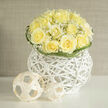 Creamy yellow roses form the heart of this carefully composed bouquet, their ruffled centres unfurling like soft sighs and gathering light within each petal. Wisps of delicate baby's breath-gypsophila-veil the roses in a froth of tiny white stars, adding air and a whispering texture that contrasts with the solidity of the blooms. The flowers are nestled in a white woven orb of rattan that reads as both vessel and sculpture: its geometric openness allows glimpses of stems and water while giving the arrangement a modern architectural base. A few minimalist woven spheres sit to one side as a contemporary accent, enhancing the composition's artful restraint. Curved blades of greenery ripple around the bouquet's edge, their subtle movement providing a gentle frame and a hint of green relief. Fragrance is understated but present-the sweet clarity of roses enriched by the clean, almost cottony scent of gypsophila and a trace of fresh earthiness like a cut meadow, conjuring early-summer mornings by Canonbury Square. Soft natural light, typical of the side streets off Essex Road, brings out the roses' buttery tones and the basket's textured white finish, making the whole piece feel airy and composed. This arrangement suits a thoughtful gift-an expression of thanks, a calming centrepiece for a small gathering, or a refined choice for a bridal detail. Crafted with the attentive balance and restraint of a local workshop, this bouquet honours both traditional charm and contemporary design, reflecting the quieter elegance often requested by customers around Canonbury. Canonbury Flowers prepares pieces like this with care for local deliveries and special moments.