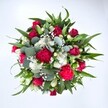 A richly detailed, top-down photograph shows a circular bouquet set against a spotless white background, allowing every bloom and leaf to stand out with clarity. At the centre, deep crimson red roses anchor the design, some still in tight bud, others just beginning to unfurl their velvety petals, suggesting a quiet, romantic promise. Clusters of fuchsia and ruby red carnations surround them, their ruffled, full heads giving the arrangement a plush, almost tactile texture that feels festive and generous. Threaded between these bold reds are delicate sprays of tiny white chrysanthemums or daisy-like flowers, each small petal radiating from a bright centre, adding lightness and a cheerful, uncomplicated charm. Fine clouds of white gypsophila, or baby's breath, drift through the bouquet, creating a soft, misty veil that seems to hover between the bolder shapes. All of this colour is cradled by layered greenery: broad, pointed dark green leaves, likely from lilies or alstroemeria, reach outward to form a strong frame, while rounded eucalyptus leaves in a silvery green weave through the gaps, lending a cool, contemporary note and hinting at their clean, herbal fragrance. The composition feels full and carefully balanced, like something just arranged on a workbench in a Canonbury flower shop, ready to travel along nearby streets towards a townhouse door or a cosy flat above a café. The bouquet's rich reds and crisp whites make it ideal for marking anniversaries, Valentine's Day, winter celebrations along Canonbury Road, or simply sending a vivid message of love and appreciation that brightens a room with its fresh, luxurious presence and heartfelt energy.
