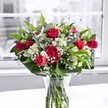 A lavish bouquet of deep red roses and ruffled carnations, gathered with white alstroemeria and clouds of delicate gypsophila, stands proudly in a clear glass vase on a bright white tabletop. The roses are the first thing the eye is drawn to: velvety crimson blooms fully open beside elegant buds, their petals layered like soft satin and suggesting a rich, romantic fragrance. Nestled among them, crimson carnations add generous volume and texture, each frilled petal catching the soft daylight that filters through a large, out-of-focus window behind. Between the bold reds, clusters of pristine white alstroemeria offer contrast, some still tightly closed, others just beginning to reveal their trumpet-shaped blossoms with faint green-tipped petals, hinting at the promise of days of beauty to come. Sprays of baby's breath weave through the arrangement like a fine mist, tiny white flowers floating against a backdrop of varied greenery. Broad, glossy deep green leaves form a natural collar at the base, while slimmer, lighter green stems-reminiscent of eucalyptus-thread upward, adding height, movement, and a quiet woodland freshness. The stems are neatly trimmed and visible through the vase, submerged in clear water that suggests the bouquet has just been prepared, perhaps for same-day delivery to a home near Canonbury Station or along Islington's leafy streets. The white tabletop reflects a soft glow back into the arrangement, and beyond the window the outdoor scene fades into a gentle, grey-toned blur, ensuring all attention stays on the vibrant reds, pure whites, and lush greens. The overall feeling is one of joyful celebration and heartfelt affection, a design that could easily mark an anniversary, a romantic gesture, or a simple but meaningful thank-you in Canonbury.