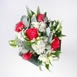 A lush hand-tied bouquet in vivid reds, pinks, and creamy whites rests against a softly lit neutral background, as if just set down on a polished Canonbury kitchen table after a walk back from Newington Green. At the heart of the arrangement, velvety crimson roses open generously, their spiralled petals catching the light and drawing the eye into rich pools of colour. Around them, pink-red carnations with ruffled, almost feathery edges add movement and texture, their layered petals giving a sense of playfulness and depth. Nestled between these bolder blooms are snow-white chrysanthemums and tiny daisy-like flowers, their clean, bright centres suggesting the clarity of a fresh morning. Wisps of delicate gypsophila float through the bouquet like a soft mist, scattering small white stars among the larger flowers and lending a feeling of airiness. Eucalyptus leaves arc outward in cool silvery-green tones, the round leaves and occasional pointed sprigs providing a modern, sculptural frame that keeps the bouquet from feeling too traditional. Behind and beneath, darker tropical greenery creates a deep, glossy backdrop that makes each bloom appear more vivid. The bouquet feels balanced yet relaxed, like something brought home to mark the start of a new chapter: a birthday, a reunion, or a quiet celebration after good news. One can almost imagine its gentle fragrance filling a living room overlooking Highbury Fields, a comforting presence that brightens the space without overwhelming it. Every stem appears thoughtfully placed, reflecting the steady, confident hand of an experienced Canonbury florist who understands how to capture both joy and calm in a single arrangement.