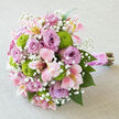 A romantic bridal bouquet lies elegantly across a light grey, linen-like fabric, its muted background highlighting every petal and stem with quiet clarity. The flowers are gathered into a generous, rounded cluster dominated by cool lavender roses, each bloom fully unfurled, with layered petals that create a sumptuous, almost velvet texture. Woven between these roses are pink alstroemeria, their trumpet-shaped petals flushed in soft pink and cream, with strokes of buttery yellow and delicate burgundy freckles that draw the eye inward. Popping brightly from the arrangement, chartreuse green button chrysanthemums form tidy, spherical mounds, their crisp, almost playful texture adding depth and a modern note to the otherwise classic palette. Scattered throughout, tiny white blossoms of baby's breath bring a gossamer lightness, as if a gentle cloud has settled over the bouquet, softening its edges. The stems are bound carefully with a wide, silky pink satin ribbon that gleams subtly in the light, and a fine strand of pearly beads spirals around the handle like a piece of bridal jewellery. Below the ribbon, the natural stems are left exposed, offering a rustic contrast to the polished finish above. The bouquet feels perfectly suited to a Canonbury wedding, perhaps carried along the leafy streets near Canonbury Square or into an intimate ceremony space off Upper Street, and would be at home in engagement photos or anniversary celebrations alike. It captures a sense of freshness and new beginnings, as if prepared early that morning by an experienced local florist and made ready for same-day delivery across Canonbury and neighbouring Islington.