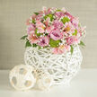 In this close-up image, a domed floral arrangement appears almost like a soft pastel cloud, carefully composed on a smooth white surface that reflects the gentle light of an overcast Canonbury afternoon. The bouquet is richly filled with roses in full bloom, their petals unfurling from the centre in tones of delicate lavender, blush pink, and deeper rose-pink that hint at romance and celebration. Nestled among the roses are pale pink alstroemeria, each flower displaying the distinctive brushstrokes and speckles along its petals, giving a sense of movement and personality. Clusters of lime green button chrysanthemums provide a lively counterpoint, their round, compact forms and vivid colour adding a playful energy and a thoroughly modern edge to the arrangement. Wisps of tiny white baby's breath float between the larger blooms, creating a fine, lacy texture that softens the overall design. A few glimpses of deep green foliage peek from beneath the flowers, adding structure and contrast without ever overwhelming the pastel palette. All of this is held within a striking white woven vase, shaped like a globe and crafted from slender wicker or rattan strands that criss-cross into an open lattice. The sculptural vase sits on the white tabletop like a piece of contemporary art, while two smaller, paler woven spheres rest alongside it, echoing the natural materials and rounded shapes. Behind them, a neutral, light beige wall keeps the composition calm and focused. The overall effect is refined yet inviting, like a wedding centerpiece ready to grace a Canonbury reception or a romantic dinner near Upper Street.