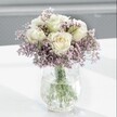 A close-up photograph shows a tranquil bouquet of creamy white roses resting in a cloud of lavender-purple statice, arranged with the quiet elegance you might expect from a Canonbury florist. Each rose sits at the heart of the design, its petals smooth and velvety, unfolding in soft spirals that reveal a gentle hint of pale yellow at the centre, as if just opening in the cool light of morning along the leafy streets off Upper Street. Around them, countless tiny blossoms of statice form an airy halo, their papery petals creating a mist-like haze of lilac and lavender tones that softens every edge and adds a gentle countryside charm. The stems are gathered in a clear glass vase with a distinctive crackled, ice-like texture that catches the natural daylight and scatters it across the smooth white surface beneath, almost like the shimmer of sunlight on Regent's Canal nearby. Through the pristine water inside the vase, the straight green stems are clearly visible, providing a fresh, grounding contrast to the pale blooms above. The background blurs into soft grey and white, with no distractions, so the viewer's gaze rests entirely on the calm beauty of the flowers. The overall feeling is one of understated serenity, the kind of bouquet that could sit quietly on a kitchen table in a Canonbury townhouse, bring comfort to a bedside, or offer a gentle, thoughtful gesture for a birthday, a get-well wish, or a moment of quiet reflection on an overcast North London afternoon.
