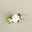 A refined boutonniere lies on a neutral, woven textile, its composition capturing the calm, understated romance of a Canonbury wedding morning. The central focus is a fully opened white lisianthus flower, its petals layered in soft, ruffled tiers that resemble fine chiffon, luminous and creamy against the simple background. Just to one side, two unopened lisianthus buds lean in gently, their pale, ivory-green forms suggesting tenderness and the unfolding of shared stories yet to come. Scattered delicately among them are sprigs of baby's breath, their clusters of tiny white blossoms creating an airy veil, like a miniature cloud encircling the main bloom and adding a sense of depth and lightness. Slim, reed-like greens and wisps of fresh foliage stretch outward from the base, introducing a subtle sense of movement, as if a breeze from nearby Canonbury Gardens had brushed past and set them in motion. The stems are gathered neatly and wrapped in a vivid pink satin ribbon, which gleams with a subtle sheen as it catches the light. The ribbon is tied into a tidy bow, its bright, cheerful tone providing a contemporary flourish that hints at personality and joy without overwhelming the gentle whites and greens. The softly textured fabric beneath offers a natural stage, highlighting every tiny petal edge and green frond. This boutonniere feels perfectly suited to be worn at an intimate registry ceremony off Upper Street or a garden celebration near New River Walk, a small but meaningful floral accent that quietly reflects care, local craftsmanship, and heartfelt celebration.