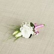 A single wedding boutonniere lies diagonally across a softly textured, light beige fabric that looks like finely woven linen, its neutral oatmeal tones echoing the calm elegance of Canonbury's quiet terraces. At the heart of the design is a fully opened white lisianthus bloom, its many ruffled petals forming a gentle, rounded cup that seems to glow against the muted background. Two creamy, pale green lisianthus buds nestle just beside it, still closed but smooth and elongated, suggesting the promise of moments on the cusp of unfolding. Tiny clouds of white gypsophila are threaded delicately throughout the arrangement, their miniature blossoms creating a gauzy halo that adds airiness and romance, like mist over nearby Highbury Fields in the early morning. Slender, grass-like blades of fresh green foliage arc gracefully from behind the central flower, extending towards the lower left, while a broader, darker green leaf sits quietly at the back, giving depth and structure. At the base, the stems are neatly gathered and wrapped in a vivid fuchsia satin ribbon, tied into a small, jaunty bow that introduces a joyful flash of colour and a slightly playful note. The ribbon's silky sheen contrasts with the more rustic weave of the fabric below, drawing the eye towards the craftsmanship of the piece. Soft, even lighting from above highlights every petal edge, leaf vein, and bead-like gypsophila flower, capturing the boutonniere as if ready to be pinned to a lapel for a Canonbury townhouse wedding, a registry ceremony near Islington Green, or a prom celebration, conveying freshness, intimacy, and a quietly modern twist on classic floral style.