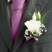 A sharply focused close-up of a groom's charcoal grey suit in Canonbury, showing a perfectly tailored jacket, patterned dress shirt, and a carefully knotted magenta silk tie, all drawing the eye towards a single, exquisite white lisianthus buttonhole. The fine wool of the suit has a subtle texture that catches the light in soft, even tones, the crisp lapel framing the boutonnière as though it were a small work of art pinned just above the heart. At the centre of the floral detail, a fully opened lisianthus bloom, creamy white with gently ruffled petals, sits like a delicate rosette, its many layers suggesting softness and quiet luxury. Just above it, two slender ivory buds lean together, not yet unfurled, hinting at new moments still to come. Around them, airy clouds of tiny white gypsophila float in every gap, their minute blossoms giving the arrangement a light, almost mist-like texture. Fine strands of linear greenery arc outward, slim and elegant, softening the transition between flower and fabric. At the base, a narrow mauve ribbon is wrapped and tied with care, its cool pink-lilac sheen echoing the deeper magenta of the tie so the whole ensemble feels thoughtfully coordinated. The scene feels like the calm pause before a ceremony in one of Canonbury's leafy churches or townhouse gardens, the fresh, green scent of the lisianthus and foliage a gentle counterpoint to polished shoes and murmured anticipation, ideal for illustrating refined wedding attire, groomsmen's styling, or special occasion flowers in this quiet corner of North London.