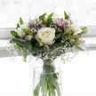 An elegant white and green bouquet stands in a simple clear glass vase, positioned against a softly glowing white background that suggests a bright Canonbury window on a still morning. At the heart of the arrangement, three creamy white roses are fully open, their velvety petals curling gently outwards to reveal ivory centres that feel fresh, cool, and perfectly formed. Around them, clusters of tiny white gypsophila, or baby's breath, drift like a light mist, their miniature blossoms creating a soft, cloud-like halo that wraps the roses in quiet tenderness. Slender stems of alstroemeria weave through the design, some still in bud and others beginning to unfurl in muted shades of mauve and blush pink, adding just a hint of colour and a suggestion of romance without disturbing the bouquet's peaceful, neutral palette. Broad, glossy green leaves fold and arc around the blooms, providing structure and an emerald frame that sets off the white flowers with natural depth and contrast. Through the cylindrical glass vase, the stems are visible, neatly gathered and tied just above the waterline with a fine ribbon, a small detail that shows the care of an attentive florist. Clear water catches the daylight, giving the whole piece a feeling of freshness, as if it has just been placed on a windowsill in a Canonbury townhouse overlooking the quiet streets near Canonbury Square. The mood is tranquil and reflective, suited to moments of remembrance, a gentle message of sympathy, or a thoughtful token of calm support delivered locally to a friend in need.