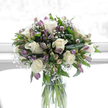 A graceful sympathy arrangement of white and purple flowers stands in a simple glass cylinder vase, bathed in soft light that recalls a calm morning in Canonbury. At its centre, smooth, creamy white roses open in generous spirals, their petals flawless and tender, conveying a sense of peace and gentle affection. They are interwoven with stems of alstroemeria whose petals shift from soft green at the base to dusky purples and cool lavenders at their tips, some blooms still in bud, others just beginning to spread their wings. The alstroemeria's elongated, slightly arching form adds rhythm and movement, as if the bouquet is breathing quietly. Fine sprays of baby's breath are scattered throughout, each tiny white flower catching the light like a dusting of frost, softening transitions between the main blooms and giving the whole design an airy, weightless quality. Deep green leaves form a supportive cradle around the flowers, their glossy surfaces and varied shapes adding natural structure and a sense of life. The clear glass vase reveals the neatly cut stems and clean water, underscoring the freshness and care that went into the arrangement, much like the meticulous attention given in a local Canonbury flower studio preparing tributes for nearby homes and services. The background is a high, bright white, without clutter or distraction, allowing the calm palette of whites, purples, and greens to stand out in quiet harmony. This bouquet feels composed with understanding and empathy, intended to bring a soft touch of beauty and reassurance to moments of loss, whether displayed in a family home or at a memorial gathering.