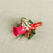 In this carefully composed boutonniere, a vibrant fuchsia rose stands proudly at the centre, its petals lush and velvety, unfolding in generous layers that catch the soft daylight like silk. The rose appears to be at the peak of its beauty, neither too tight nor overly open, giving the impression of a promise held tenderly in the moment. Surrounding it, delicate sprigs of white baby's breath form a gentle halo, each tiny blossom contributing a lace-like softness that contrasts beautifully with the richness of the rose. Clusters of scarlet hypericum berries nestle at its base, round and polished, adding a playful rhythm of colour reminiscent of the warm, lively feel of Canonbury's cafés and side streets. The composition is framed by neatly curled aspidistra leaves and smooth green foliage, shaped into loops that guide the eye around the design, while fine threads of grass introduce a sense of lightness and movement, as if ready to sway with each step down a Canonbury Place pathway. The stem is wrapped in a coordinating red ribbon, wound with measured precision, its finish smooth and slightly glossy, lending the design a tailored, almost sartorial quality. Resting against a soft, neutral textile background, the boutonniere's colours and forms stand out in calm relief, refined yet approachable. You can almost sense the cool, fresh touch of the greenery and the quiet, sweet scent of the rose, ideal for a groom, best man, or prom guest preparing in a nearby townhouse. It carries the feeling of something crafted with time, skill, and affection, a small floral artwork to be worn throughout a cherished day.