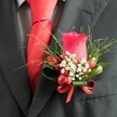 A close-up, high-angle photograph reveals an elegant boutonnière pinned precisely to the left lapel of a deep charcoal suit, the kind you might see on a groom stepping out of a townhouse near Canonbury Square. At the centre stands a single, perfectly formed deep red rosebud, its velvety petals spiralling inward in tight, confident layers, catching the soft indoor light with a gentle sheen. Around the base of the rose, a halo of tiny white gypsophila blooms, like a cloud of baby's breath, softens the intensity of the red and adds a sense of airiness and romance. Nestled among these star-like flowers are several small, glossy red berries, their rounded surfaces reflecting the light and echoing the rose's rich hue, adding both texture and a playful detail to the design. Slim, grass-like fronds of greenery fan out behind the flowers, adding movement and height, while broader, darker leaves cup the base and give structure, suggesting the careful hand of an experienced Canonbury florist. The stems are neatly bound and finished with a narrow red satin ribbon, tied into a small bow that mirrors the depth of the rose and berries. Just below the boutonnière, a sharply knotted red tie in smooth satin or silk draws the eye, reinforcing the cohesive red colour palette and giving the whole ensemble a polished, formal air. Beneath the tie, a subtly patterned dress shirt in pale grey or white, perhaps a fine check or houndstooth, peeks through, creating an additional layer of refined texture. The overall mood of the image is one of poised celebration, perfect for weddings, proms, or black-tie events across Canonbury and surrounding North London streets.