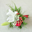 Laid gently against a soft, neutral backdrop, this hand-tied bouquet has the composed elegance of a wedding party pausing for photographs near Canonbury Square. The eye is first drawn to a luminous white lily, fully open, its silky petals clean and unblemished, forming a graceful star at the centre of the design. Within, a dusting of pale pollen adds a touch of natural warmth, evoking purity, hope, and the promise of a new chapter. Encircling this focal bloom, rich red roses unfold in tight spirals of velvety petals, their deep crimson shade suggesting passion, loyalty, and enduring affection. Bright red berries, clustered along slender stems, punctuate the arrangement with cheerful bursts of colour that feel almost like confetti tossed in celebration along nearby Essex Road. Wisps of gypsophila thread through the bouquet, their tiny white flowers creating a delicate haze that softens the stronger hues. Fine, feathery greenery stretches outward, adding shape and movement, giving the entire piece a natural, slightly whimsical silhouette without losing its sense of refinement. The stems are cleanly trimmed and bound with a smooth scarlet ribbon, snug yet understated, as if ready to be carried down the aisle at a Canonbury wedding or presented during a heartfelt engagement dinner. There is a gentle balance between drama and tenderness here-the confident contrast of red and white, the interplay of lush petals and airy foliage, the subtle fragrance that would greet you as the bouquet is lifted from its wrapping. It feels perfectly suited to marking cherished moments across Canonbury and nearby Islington, from romantic gestures to intimate family celebrations.