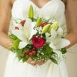 This hand-tied bridal bouquet radiates organic beauty and soft sophistication, gently resting in a bride's hands as she stands adorned in a white gown with intricate, textured details. The crisp white lilies unfurl at the heart, luminous as early morning light on Canonbury Green, while rich crimson roses pulse with a romantic energy, each velvety swirl recalling promises whispered along the tree-lined avenues nearby. Unopened green lily buds lend a fresh, hopeful spirit, complemented by frothy clouds of baby's breath that float gracefully throughout the arrangement. Pops of festive red from hypericum berries accent the bouquet, their cheerful glow set off by spherical moss-green elements reminiscent of dewy lawns on New River Walk. Slender and broad greenery, gathered with an artist's care, creates luxurious depth and textural charm, as if the flowers have just been gathered from a local garden at dawn. Set against a pale, softly blurred backdrop, the bouquet's intricate forms and classic palette evoke timeless devotion and quiet celebration, perfect for weddings or any moment when love-and Canonbury's own gracious elegance-takes centre stage.