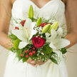 Cradled against the delicate lace of a bridal gown, this hand-tied bouquet looks like a moment of Canonbury romance captured in flowers. At its heart, a single red rose glows with velvety intensity, drawing the eye with the richness of deep wine, while three or so white lilies unfurl around it in serene, star-shaped blooms. Their creamy petals appear almost luminous against the soft fabric of the dress, hinting at the gentle fragrance that seems to hover in the still air, like the scent of blossoms along Canonbury Grove on a warm day. Between the larger flowers, sprigs of gypsophila and baby's breath scatter tiny white dots that soften the composition, adding a cloud-like lightness and echoing the intricacy of the bride's lace. Ruby-red berries peek through the foliage, lending a joyful spark reminiscent of winter weddings or festive gatherings in local Islington churches. Textured mossy accents and fresh green leaves layer different shades of green, from bright new shoots to deeper forest tones, giving the bouquet a natural, slightly wild charm. Slender grasses arc outward, creating an almost floating effect, so the bouquet feels alive and gently in motion, as if it has just been lifted from a nearby Canonbury garden. The arrangement is tightly composed yet never stiff, radiating a sense of poised celebration and quiet luxury. It's the kind of bouquet a local florist would create for a bride who wants tradition warmed by personality, a piece that will be remembered in photographs and in memory long after the last petal has fallen.