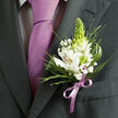 This image offers an intimate, waist-up glimpse of formal menswear, composed with the same care a florist lavishes on a bridal bouquet. The focus rests on the left lapel of a dark charcoal grey suit jacket, its fabric smooth and well-cut, suggesting a special occasion unfolding somewhere around Canonbury's leafy streets. Beneath the jacket, a light-toned dress shirt with a discreet dark micro-check pattern adds quiet interest without stealing the show. The eye is drawn to a sleek lavender tie knotted neatly at the collar, its satin surface reflecting soft, diffused light like dusk along the New River Walk. Just beside it, pinned precisely to the lapel, a delicate boutonniere becomes the visual centrepiece. White alstroemeria-Peruvian lilies-open in gentle curves, each petal detailed with fine reddish-brown brushstrokes and warm yellow centres. They cradle a striking, upright green bud, tall and tapering, whose fresh lime hue brings a subtle modern edge. Surrounding this focal point, a cloud of white gypsophila dots the arrangement with tiny blooms, while wisps of ornamental grass and a mix of narrow spikes and broader, polished green leaves provide movement and depth. The stems are gathered and finished with a lilac satin ribbon, tied in a neat bow that echoes the colour of the tie and reinforces the sense of coordination. The composition feels cool, clean, and hopeful-like the start of a wedding at a hidden Canonbury garden or a prom night beginning with a photograph on a quiet Islington terrace-capturing the quiet confidence of someone dressed with intention, right down to the last petal.