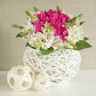 This image captures a small yet sumptuous bridal arrangement that feels perfectly at home in a Canonbury townhouse reception. At the centre, vivid fuchsia blooms-full, petal-rich and reminiscent of peonies in high season-are gathered tightly into a glowing cushion of colour, their velvety texture drawing the eye and inviting the fingertips. Encircling this bold heart, soft ivory and white alstroemeria unfold in layers, each flower gently streaked with hints of blush and pale lime, creating a lively interplay of light and shadow. Slim green stems and unopened buds rise carefully between the blossoms, adding height and movement, like the first shoots in Arlington Square after spring rain. The flowers sit snugly inside a round white wicker basket, its crisp, painted finish and intricate interwoven bands giving it a sculptural, modern feel. Two small cream-toned decorative orbs, also woven, rest close by on a clean white surface, subtly mirroring the basket's texture and bringing a contemplative calm to the arrangement. The lighting is soft and filtered, as if coming through a sheer curtain on a quiet Canonbury afternoon, illuminating each petal edge and making the greens glow with freshness. There is a sense of thoughtful luxury here, suitable for a wedding breakfast table, a signing table at Islington Town Hall, or a romantic post-ceremony dinner at home. The imagined fragrance is light and green, calming yet uplifting. Crafted with an eye for both structure and emotion, this bouquet speaks of new beginnings, celebration, and the gentle sophistication that Canonbury Flowers brings to wedding deliveries throughout N1.