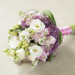 A meticulously hand-tied bouquet lies horizontally on a pale beige fabric with a subtle weave, the kind of gentle texture that feels at home on a Canonbury dining table or windowsill overlooking the leafy streets. The arrangement is built around generous white lisianthus blooms, some fully open with soft, layered petals cupping a hint of yellow-green at the centre, others still enclosed in tight, elegant buds that add freshness and promise. Interlaced with these are tall stems of stock in shades of blush pink and light mauve, their clusters of ruffled florets creating a billowing, cloudlike effect that softens the entire design. Nestled among the pastels, clusters of small, star-shaped purple flowers, reminiscent of ornamental allium, punctuate the bouquet with a bold lavender-purple accent and a distinctive, slightly spiky texture. Long, slender strands of deep green foliage arc and loop through the blooms, particularly along the right edge, guiding the eye and lending a graceful rhythm to the composition. The gathered stems are secured with a vivid fuchsia satin ribbon, wrapped smoothly and finished with a fine string of tiny white pearls, which glint subtly as if catching late afternoon light along nearby Canonbury Square. The bouquet feels elegant and composed yet tender, suitable as a bridal bouquet, a special anniversary gift, or a centrepiece for an intimate celebration. You can almost sense the gentle perfume of stock and the crisp, green scent of foliage, a carefully balanced arrangement that speaks of craftsmanship, romance, and thoughtful design, perfect for same-day delivery across this quiet corner of North London.