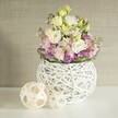 A celebration of handmade elegance, this arrangement sits at the heart of a bright Canonbury home, reflecting the artistic essence of the neighbourhood's leafy crescents and garden studios. The centrepiece is a rounded, modern vase, its openwork weave of white rattan giving it both lightness and sculptural character, reminiscent of the creative textures found in nearby Camden Passage shops. Overflowing from the vase are soft, ruffled lisianthus blooms in shades of ivory and pastel yellow, their gentle forms interspersed with slender stalks of pink matthiola, offering vertical movement and a sense of lush abundance. At the bouquet's base, lively purple filler flowers-perhaps asters or little alliums-add richness and visual depth, bordered by a collar of glossy green foliage that frames the entire display. Two decorative spheres, crafted from pale, woven rattan, rest on the left of the pristine table, subtly uniting the composition and suggesting a love for handcrafted detail. Bathed in natural, diffused light and backed by a warm beige textured wall, the arrangement creates a mood of calm sophistication-ideal for a wedding breakfast on Canonbury Road, a creative brunch, or simply inviting the spirit of spring into a North London living space. This harmonious combination invites feelings of freshness, comfort, and artistry that's true to Canonbury's local style.