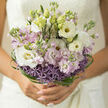 This bouquet appears as a piece of living poetry, the sort of carefully balanced design you might see carried from a townhouse doorway in Canonbury towards a ceremony in Islington. A gentle cloud of pastel lilac and creamy white blooms is gathered into the hands, featuring layers of lisianthus and stock whose petals unfurl like pages of a love letter. Their soft, velvety surfaces catch the daylight, giving the impression that each flower is quietly glowing, touched by the calm brightness of an early afternoon near Canonbury Square. At the heart and base of the bouquet, dusky lavender allium forms a ring of tiny star-shaped florets, creating a subtle yet striking contrast in both form and depth of colour. The purples peek out from beneath the paler blossoms like hidden sparks of joy, hinting at celebration without overwhelming the tranquil palette. Around the edge, glossy strands of green bear grass curl and loop in precise spirals, framing the flowers with an almost sculptural outline that showcases the florist's meticulous hand-tied technique. Fresh green buds are interspersed throughout, offering a sense of anticipation, as though more beauty is still to come. The bouquet feels light and airy at the top, with blooms placed to breathe and move gently, while the base is more compact, giving stability in the hands of a bride walking along the tree-lined streets around St Paul's Road. You can almost catch a trace of fragrance in the air: the sweetness of stocks mixing with the gentle, clean scent of lisianthus, evoking quiet happiness and hope-filled moments. This is a bouquet that would feel perfectly at home at an elegant Canonbury wedding, a renewal of vows, or a cherished anniversary, its understated sophistication allowing the emotion of the day to shine through.