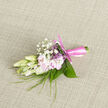 Resting on a softly woven, neutral cloth, this refined boutonnière appears as though it has just been set down for a final check before being pinned onto a suit in a Canonbury townhouse near the New River Walk. At first glance, your attention is drawn to the creamy buds at the centre, their petals firm yet tender, with a luminous quality that suggests they will open fully later in the day. Tucked beside them are pale blush-pink blooms, their petals loose and airy, curling gently at the edges with the softness of a sigh. Interspersed among these are delicate sprigs of baby's breath, each tiny white flower like a miniature cloud floating above the main blossoms, adding texture without overwhelming the composition. Narrow, blade-like strands of greenery sweep gracefully through the flowers, while slightly broader leaves create a neat, natural frame around the base. The stems are carefully aligned and bound in a satiny pink ribbon, its smooth surface catching a hint of the ambient light, the bow tied with the kind of precision that speaks of an experienced florist's hand. There's an impression of a light, sweet fragrance, subtle yet evocative of freshly cut bouquets being prepared for local weddings and celebrations. The overall palette of soft ivory, blush pink, and fresh green feels modern but timeless, perfectly suited to a groom, usher, or prom guest walking through Canonbury towards an Islington venue. This boutonnière balances romance and restraint, bringing a gentle note of nature, craftsmanship, and personal detail to any special day.