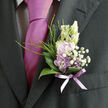 Pinned neatly to the lapel of a dark, finely tailored suit, this boutonnière offers a soft whisper of colour that feels perfectly at home in an elegant Canonbury townhouse wedding. A cluster of pale lavender blossoms forms the heart of the design, their petals smooth and slightly translucent, curling gently at the tips as though just opening. They are framed by tiny white filler flowers, most likely gypsophila, whose many small, star-like heads give a delicate, clouded texture, catching the light like tiny droplets of morning mist. Around them, a collar of glossy green leaves curves protectively, each leaf broad and gently arched, adding depth and a sense of natural structure reminiscent of mature shrubs along the quiet streets near Canonbury Square. Sprigs of slender, grassy foliage peek through, lending a subtle wildness, as though these stems had been selected from the edge of a meadow rather than a shop display. At the base, a sheer lavender ribbon is wrapped with meticulous care, its slightly translucent fabric allowing hints of the green stems to show through, and the soft purple tone gently echoing both the flowers and the nearby tie. Against the deep charcoal of the suit fabric, the colours appear even more serene and romantic, drawing the eye without shouting. The overall impression is one of thoughtful understatement: a floral accent that carries the scent and freshness of early spring, perfectly suited to a wedding or formal gathering in Canonbury, where refined style and a relaxed, neighbourhood warmth often sit side by side, much like the blooms and greenery in this small yet meaningful arrangement.