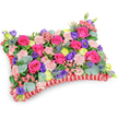 A vibrant, pillow-shaped floral tribute sits photographed from a gently elevated, oblique angle on a clean white background, its softened edges and dense surface clearly visible. The top of the cushion is a lush carpet of blooms: velvety hot-pink roses dominate, their cupped petals ranging from tight buds to partially open blooms that catch the light like small lanterns. Scattered among them are pale shell-pink carnations with their frilled edges, and deep purple lisianthus that add graceful, bell-shaped contrast. Bright lemon-yellow ranunculus punctuate the composition with sunny, buttercup-like blooms, while a few silver-blue eryngium thistles lend a dramatic, spiky architecture and a cool tonal foil to the warmer palette. Rounded, silvery-green eucalyptus leaves and varied glossy greens weave through the flowers, giving depth and a soothing herbal scent that balances the sweetness of the roses. Around the perimeter a rich, deep red pleated satin ribbon has been ruffled and secured to form the classic cushion border, its soft sheen framing the floral tableau and defining the rectangular pillow silhouette. Soft, diffused studio lighting emphasizes texture and colour without harsh shadows, allowing each petal and leaf to read clearly against the white backdrop. As a sympathy or memorial cushion the arrangement reads as a celebration of life rather than solemn mourning: the mixture of fuchsia, lavender, lemon and blush feels comforting and warm. Made with attentive craftsmanship known in Canonbury, this pillow tribute evokes quiet solace - a fragrant, tactile reminder of affection suited for services near Canonbury Square or a peaceful graveside moment.