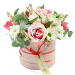 A charming pastel hatbox bouquet is shown centred against a clear white background, the simplicity of the setting allowing the soft pinks, whites, and greens to glow with quiet elegance. The box is a cylindrical, pale pink hatbox with a smooth matte texture, reminiscent of stylish gift packaging you might see in independent Canonbury boutiques. Around its base, a narrow coral-red satin ribbon is tied into a neat bow, the sheen of the fabric catching the light. From this bow hangs a small, cream tag with gentle green calligraphy-style text, hinting at a personal message or local delivery. The flowers spill generously over the top in a full, rounded dome, arranged with the care of an experienced florist. A prominent, bi-colour rose in pale pink with a more intense pink centre takes pride of place, its open, velvety petals creating a romantic focal point. Surrounding it are fresh white chrysanthemums with vivid green centres, each bloom crisp and daisy-like, adding lightness and clarity. Slender stems of white alstroemeria, speckled softly on the inner petals, weave through the design, joined by clusters of deep pink flowers-likely spray roses or lisianthus-that provide rich, playful bursts of colour. The bouquet is framed with varying foliage: silver-green eucalyptus leaves in rounded shapes that suggest a cool fragrance, deeper green leaves that ground the palette, and delicate sprays bearing tiny green buds that speak of new beginnings. The overall look feels like a gentle celebration, suited to birthdays, new home gifts, or quiet romantic gestures, and could easily be pictured arriving by same-day delivery to homes along Canonbury Road, Essex Road, or the leafy streets leading towards Islington Green.