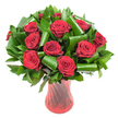 A lush, rounded bouquet of a dozen or more deep crimson red roses fills the frame, each bloom open and velvety, petals spiralling from tight centres into flawless, satiny layers. The roses are densely clustered to create an opulent dome of colour - a wine-dark red that reads both passionate and refined under soft, even studio light. Interwoven throughout the heads of the roses is abundant dark green foliage: glossy, flat leaves that form a verdant backdrop, and broader, emerald-green leaves artfully rolled and curved into sculptural loops and cylinders. Those folded leaves add a contemporary, architectural rhythm against the gentle softness of the petals, their polished sheen catching the light and framing each rose like a protective collar. The arrangement is anchored in a tall, translucent ruby-red glass vase with a subtle ribbed or twisted texture; its vertical lines echo the stems and catch fleeting reflections, visually deepening the bouquet's ruby tones. Set against a clean white background, every detail - the velvety petal edges, the curled tips of the foliage, the glass's faint swirls - reads with clarity, drawing the eye to texture and craft. The overall impression is classic romance with a modern twist: a bouquet that suggests anniversaries, Valentine's Day, or an intimate celebration, redolent of a soft, familiar rose scent that seems to hang in the air. For someone in Canonbury or nearby Newington Green seeking a statement of enduring love, this arrangement offers both timeless symbolism and contemporary floral artistry.