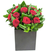 This modern bouquet centres on a compact cluster of rich, velvety red roses whose petals open in soft, generous spirals, each bloom holding a deep, almost satin crimson that catches the light like small red lamps. They sit tightly grouped so the eye reads a single, passionate heart; around them, broad glossy green leaves have been carefully curled and layered to form rhythmic arcs that both cradle and propel the roses upward, adding sculptural movement and a cool, tropical clarity. The flowers are arranged in a low, matte black cube that gives the composition a grounded, architectural feel - the contrast between the luminous petals and the vase's flat surface heightens the visual drama. Lighting is bright and contemporary, creating soft highlights on leaf edges and a gentle sheen across every petal so that the bouquet reads as both intimate and polished. Close to the arrangement you'd imagine the clean scent of fresh greenery mingling with the classic, heady perfume of roses; the aroma is restrained rather than overwhelming, a reminder of careful handling and freshness delivered same day across Canonbury and nearby Islington streets. As a local florist would say, this design balances passion and restraint - ideal for anniversaries, a romantic apology, or to bring an elegant, heartfelt presence to a modern home or office on Essex Road. The overall effect is confident, tactile, and quietly luxurious: an expression of deep feeling shaped with expert, neighbourhood care.