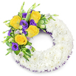 A reverent, circular sympathy wreath fashioned from a plush ring of small, pristine white spray chrysanthemums, photographed against a soft white backdrop. The chrysanthemum cluster is densely packed to create a cloud-like, cushiony texture; each tiny, layered petal overlaps in a way that reads as calm and continuous, a comforting visual hush. Nestled on the upper left curve is a single, deliberate spray of colour: several fully opened yellow roses with buttery, spiralled petals that suggest warmth and gentle remembrance, set beside deep purple lisianthus (Eustoma) whose ruffled, cup-shaped blooms offer a velvety contrast. A few lisianthus buds in palest lavender and green add points of quiet growth, while varied green foliage - glossy salal-like leaves, a few variegated touches and delicate fern fronds - fans beneath the flowers to give shape and natural movement. A discreet dark purple mesh or crinoline ribbon peeks from beneath the chrysanthemum edge, echoing the lisianthus and adding a slender, refined line around the inner and outer rings. The whole composition sits with soft, even lighting that preserves the whites' purity and lets the yellow and purple sing without overpowering the wreath's solemn intent. As a local florist I picture this piece delivered to services around Canonbury Square or laid by the memorial table at a chapel on Essex Road: it reads as both respectful and tender, a carefully balanced tribute that speaks of peace, memory and the understated grace of close community remembrances in Canonbury.