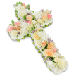 A large, reverent floral cross lies against a clean white background, its shape completely covered in a soothing blend of white and pastel flowers that evoke quiet sympathy and faith. The cross itself is full and rounded, with no gaps visible, built from layers of tightly packed white and creamy blooms that give it a dense, luxurious texture. At intervals along the length and arms of the cross, clusters of roses in soft peach, apricot, and the palest blush pink rise slightly above the white base, drawing subtle lines of colour that guide the gaze and add gentle warmth. Nestled between these feature roses are numerous white hydrangea petals and small white spray roses or similar blossoms, so that every inch appears richly decorated. Small touches of lavender-hued filler flowers punctuate the arrangement, bringing tiny, carefully placed notes of pale purple that add intricacy and visual interest. Fine sprigs of fresh greenery can be seen peeking at the edges, tracing the outline of the cross and providing contrast against both the flowers and the bright background. The lighting is even and natural, highlighting the softness of the petals and the thoughtful craftsmanship behind the piece. This floral cross feels like a deeply respectful tribute that would be placed near the coffin or at the front of a chapel in Canonbury, offering comfort to mourners and symbolising hope, eternal life, and the enduring love that surrounds a farewell service.