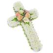 A refined floral sympathy cross dominates the frame, standing out clearly against a bright white background that enhances every detail of the design. The cross itself is constructed from a dense covering of white button chrysanthemums, their small, rounded flower heads packed so tightly that they form a plush, continuous surface over the entire structure. This blanket of white feels calming and pure, evoking silence and peace, like an early morning in Canonbury before the streets fully stir. Running along the outer edge of the cross, just visible beneath the thick covering of blooms, is a slim band of lime green ribbon. This simple but effective trim outlines the shape, adding a fresh, contemporary touch and a hint of lively colour without taking away from the overall solemn mood. At the junction where the vertical and horizontal beams meet, a carefully arranged cluster of roses becomes the visual and emotional heart of the piece. The roses open fully in soft shades of blush pink, creamy ivory, and warm peach, their velvety petals layered in graceful spirals. Between them, fine fern fronds add feathery greenery, while tiny sprigs of light lavender or purple filler flowers introduce delicate accents of colour. The cross exudes quiet elegance and reverence, making it a fitting floral tribute for a funeral, church service, or memorial held in Canonbury or along neighbouring Upper Street. It conveys sympathy, love, and faith in a way that feels both traditional and gently contemporary. The craftsmanship suggests the steady hands of an experienced local florist, creating a comforting symbol of remembrance and hope for grieving families.