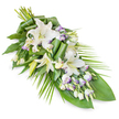 A dignified, elongated sympathy spray of white lilies and lisianthus is displayed diagonally on a bright white background, the composition carefully shaped so it appears to flow gently from a fuller, rounded head at the upper left down to a narrow, pointed tip at the lower right. Several large oriental lilies form the focal point, their pristine white, trumpet-shaped petals unfolding gracefully, while part-closed buds rise among them like gentle promises of remembrance. Hints of pale yellow-green appear in the stamens, drawing the eye to the centre of each bloom without disturbing the cool, calm mood. Around and between the lilies, clusters of lisianthus offer smaller, more delicate flowers and slender buds; their soft white petals are touched at the edges with faint lavender, a nod to the misty tones often seen in early morning skies over nearby New River Walk. The arrangement rests on a foundation of mixed greenery: broad, glossy leaves give structure and weight, while finer, palm-like fronds arch outward from the right, helping create the graceful, asymmetric silhouette. A few leaves are rolled and sculpted into smooth curves, adding a contemporary feel while keeping the tribute understated and respectful. The spray feels soothing and composed, like a quiet corner of Canonbury where families gather to share memories. Ideal for placing on a casket, beside a memorial book, or at a crematorium chapel, this floral piece visually expresses sympathy, serenity, and enduring love, offering a gentle, natural presence at final farewells in Canonbury and surrounding North London neighbourhoods.