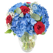 A luminous, hand-arranged bouquet sits in a clear glass vase, its rounded silhouette filled with water and neatly visible stems. The composition is dominated by voluminous clusters of sky-blue hydrangeas that form a soft, cloud-like mass; each tiny floret overlaps like ripples of summer sky after a rain. Nestled through that blue expanse are three deep, velvety red roses-tightly furled at the centre, their petals rich and sensual-providing a dramatic, romantic punctuation against the cool hydrangea tones. Sprays of delicate white baby's breath float between the larger blooms, lending an airy, starry softness that brightens the overall texture. Muted, rounded eucalyptus leaves and fresh green foliage weave through the arrangement, their silvery-green tone adding quiet herbal depth and a faint, uplifting scent when the bouquet is handled. The scene is lit with bright, clean light against a white backdrop, which highlights the saturated blues and reds and the sheen on the rose petals, while the clear vase reveals the crisp freshness of the stems below. As an experienced Canonbury florist might say, this arrangement balances bold colour with gentle texture-perfect as a centerpiece for a celebration on a sunlit dining table, an anniversary gift that speaks of devotion, or a patriotic display for seasonal gatherings. The bouquet feels confident yet soothing, designed to be both visually striking and quietly fragrant, a hand-crafted statement that lifts a room and honours moments of joy, remembrance, and connection in the local neighbourhood.